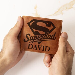 May include: A brown leather wallet with the words "Superdad" and "DAVID" printed on the front. The design includes a stylized "S" within a diamond shape. The wallet is held in two hands.