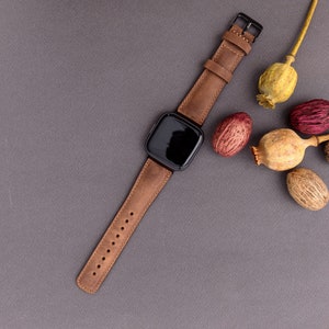 May include: A brown leather watch band with a black square watch face. The watch is on a gray surface with dried poppy seed pods.