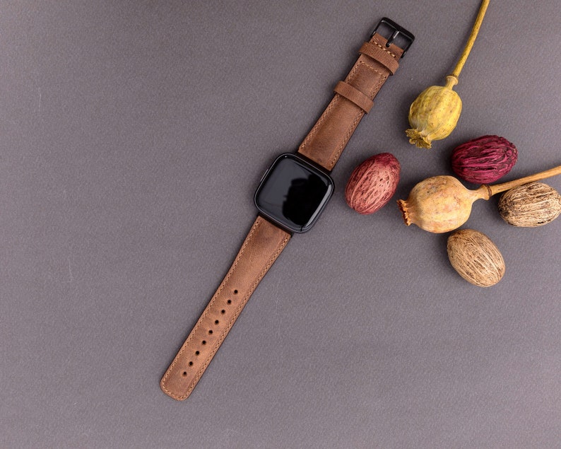 May include: A brown leather watch band with a black square watch face. The watch is on a gray surface with dried poppy seed pods.