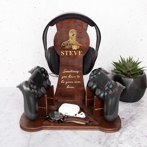 May include: A wooden gaming console stand with a personalized name "STEVE" and a quote "Sometimes you have to be your own hero." The stand has slots for two controllers, a headset, and a phone.
