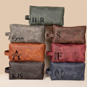 May include: A collection of personalized leather toiletry bags in various colors, including gray, brown, and navy. Each bag features a unique monogram or name in black lettering. The bags have a rectangular shape with a zipper closure and a side handle.