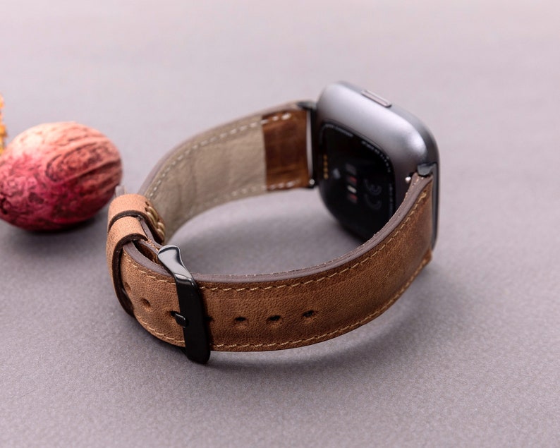 May include: A brown leather watch band with a black buckle. The band is designed for a smartwatch with a square face.