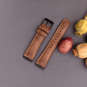 May include: Two brown leather watch straps with black metal buckles. The straps are laid out on a gray surface with dried poppy seed pods.