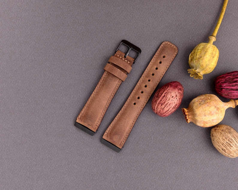 May include: Two brown leather watch straps with black metal buckles. The straps are laid out on a gray surface with dried poppy seed pods.