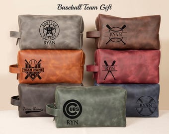 May include: Six different leather toiletry bags with baseball team logos and names. The bags are in various colors, including gray, brown, red, green, and blue. The logos include the Houston Astros, Chicago Cubs, and a generic baseball logo. The names on the bags are "Ryan" and "Team Name".