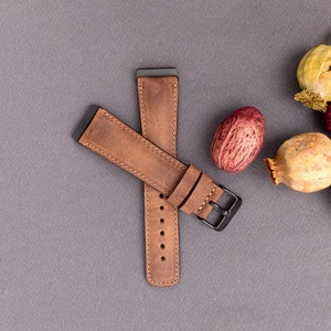 May include: A brown leather watch band with a black metal buckle. The band is laid out on a gray surface.