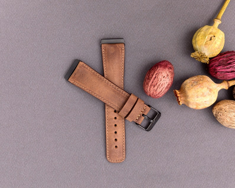 May include: A brown leather watch band with a black metal buckle. The band is laid out on a gray surface.
