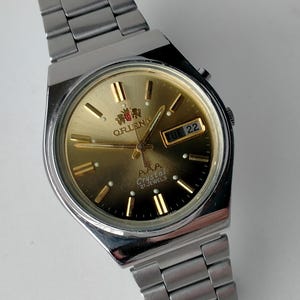 May include: A silver-toned wristwatch with a gold-colored face. The watch features a day and date display, gold-colored hour markers, and the brand name "ORIENT". The watch band is made of silver-toned metal links.