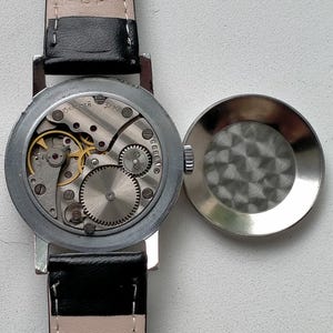 May include: An open-faced vintage wristwatch with a black leather strap. The watch mechanism is visible, showcasing intricate gears and a silver-colored casing. The watch face is open, revealing the internal components.