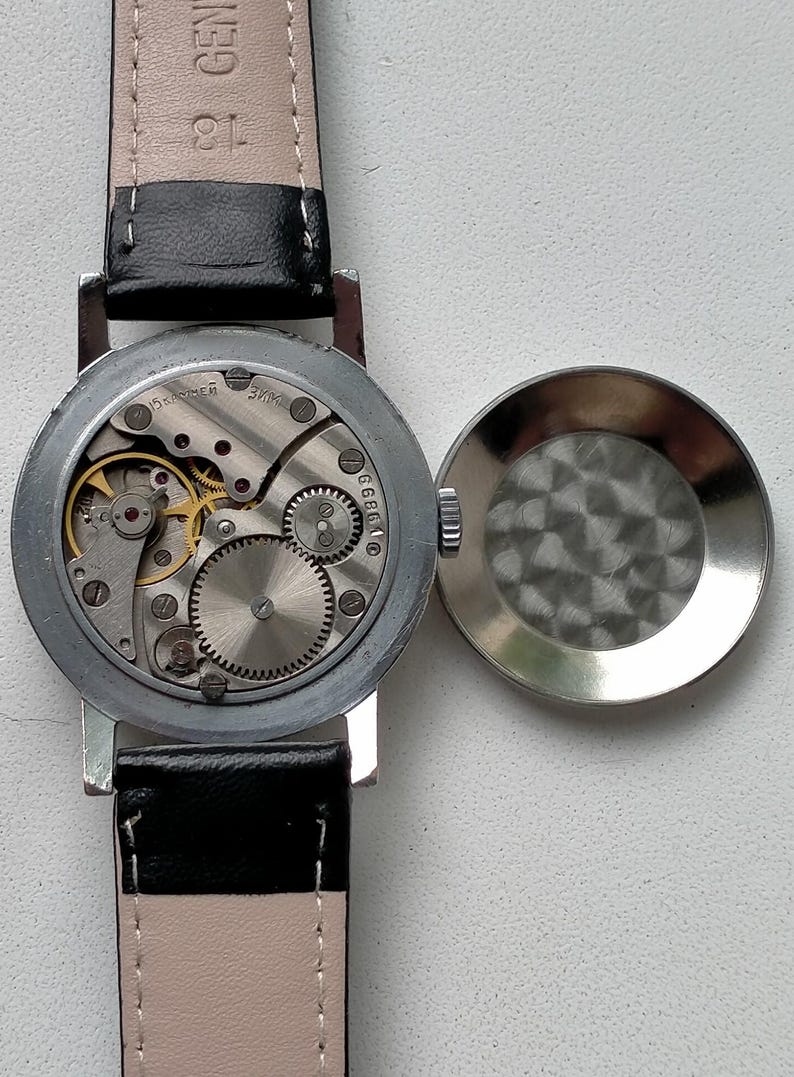 May include: An open-faced vintage wristwatch with a black leather strap. The watch mechanism is visible, showcasing intricate gears and a silver-colored casing. The watch face is open, revealing the internal components.