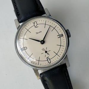 May include: A vintage wristwatch with a cream-colored face, black hands, and a black leather band. The watch face features black numerals and markings, with the word "Зим" printed on the face. The watch has a silver-colored case.
