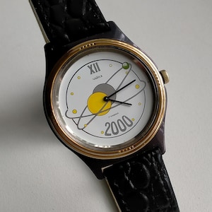 May include: A black leather wristwatch with a gold-tone bezel and a white face. The face features a stylized solar system design with the text "Чайка 2000" and "17 камней".