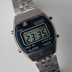 May include: Silver-toned digital watch with a rectangular face and a matching metal band. The watch face displays the time, day, and date. The watch has buttons for light, timer, alarm, and calendar functions. The brand name "Anker" is visible on the watch face.