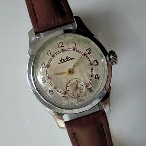 May include: Vintage wristwatch with a silver-tone case and a brown leather band. The watch face is off-white with black numerals and the brand name "Heba". The watch has a small seconds dial and red markers. A classic timepiece.