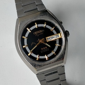 Puede incluir: Reloj de pulsera automático Carnavin Crystal plateado con esfera negra. El reloj presenta agujas e indicadores dorados, una pantalla de día y fecha y una correa de acero inoxidable. La esfera muestra "MON 26".