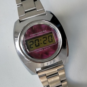 May include: A vintage silver digital wristwatch showing the time 20:20. The watch face has a pinkish-purple background with the word "Электроника" and "сделано в, СССР" printed on it. The watch band is made of metal.
