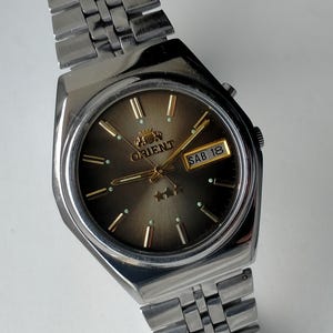 May include: A silver Orient watch with a brown face and a metal strap. The watch face displays the Orient logo, gold hands, and the day and date. The watch has a rectangular case and a silver metal strap.