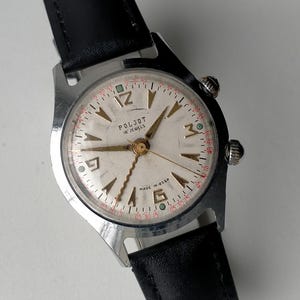 May include: Vintage Poljot 18 Jewels wristwatch with a silver-colored face, gold-tone hands, and black leather band. The watch face features the text "POLJOT 18 JEWELS" and "MADE IN USSR". The watch has a silver-colored case and two crowns.