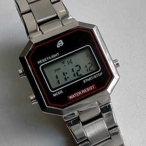 May include: A silver digital watch with a black face and red detailing. The watch shows the time 11:12:12 and has the text "WATER RESIST". The watch strap is made of silver metal.