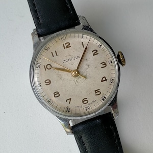 Könnte beinhalten: Vintage Armbanduhr mit silberfarbenem Gehäuse, cremefarbenem Zifferblatt und schwarzem Lederarmband. Das Zifferblatt hat goldfarbene Zeiger und Ziffern, mit dem Wort "ПОБЕДА" auf dem Zifferblatt. Durchmesser: ca. 3,5 cm.