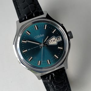 Puede incluir: Un reloj de pulsera vintage con esfera de color verde azulado y caja plateada. El reloj tiene una correa de cuero negro y una pantalla de fecha. La esfera del reloj tiene la palabra "Слава" y el texto "26 камней" impresos.