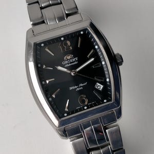 May include: A silver Orient Automatic watch with a black face. The watch has a rectangular dial with a date display, luminous hands, and a stainless steel strap. The watch face displays the text "Orient Automatic" and "Water Resist 50m".