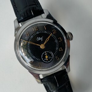 May include: A vintage wristwatch with a silver-tone metal case and a black leather strap. The black dial features gold-colored Arabic numerals, gold hands, and a small sub-dial for seconds. The Cyrillic text "свет" is printed in white on the dial.