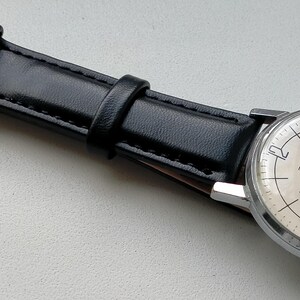 May include: A vintage wristwatch with a silver-toned case and a cream-colored face. The watch has black hands and hour markers, and a small seconds dial. It features a black leather strap with a silver buckle.