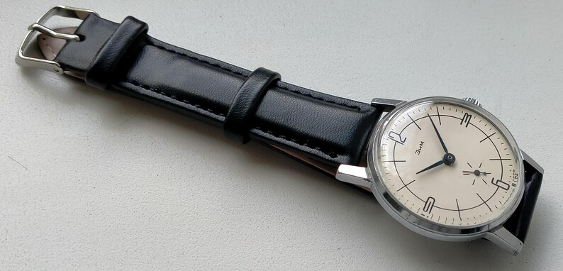 May include: A vintage wristwatch with a silver-toned case and a cream-colored face. The watch has black hands and hour markers, and a small seconds dial. It features a black leather strap with a silver buckle.