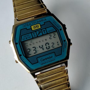 May include: A gold-tone digital wristwatch with a metallic expansion band and a rectangular case. The watch face features a bright blue background with black text including "ЭЛЕКТРОНИКА" and "Сигнал", displaying the time 23:40:21 and the date 22.