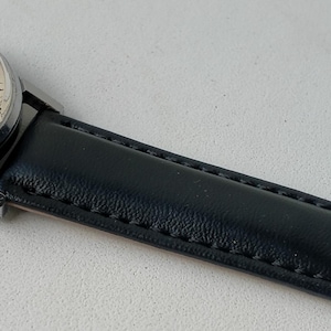 May include: A vintage wristwatch with a silver-colored case and a cream-colored face. The watch features black hands and hour markers, and a black leather band. The watch is displayed at an angle, showcasing its classic design.