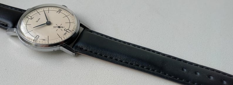 May include: A vintage wristwatch with a silver-colored case and a cream-colored face. The watch features black hands and hour markers, and a black leather band. The watch is displayed at an angle, showcasing its classic design.