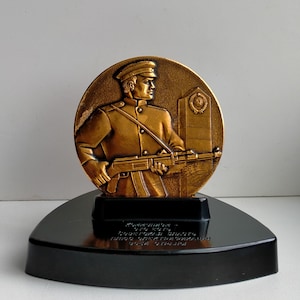 Op de afbeelding: Een bronzen medaille met een reliëf van een soldaat die een geweer vasthoudt. De medaille staat op een zwarte driehoekige voet met de inscriptie "Beloning voor de besten der besten, ter bescherming van de Sovjet-Unie".