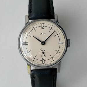 May include: A vintage wristwatch with a cream-colored face, black hands, and a black leather band. The watch has a silver-colored case and the word "Зим" is printed on the face. The watch band is approximately 0.75 inches wide.