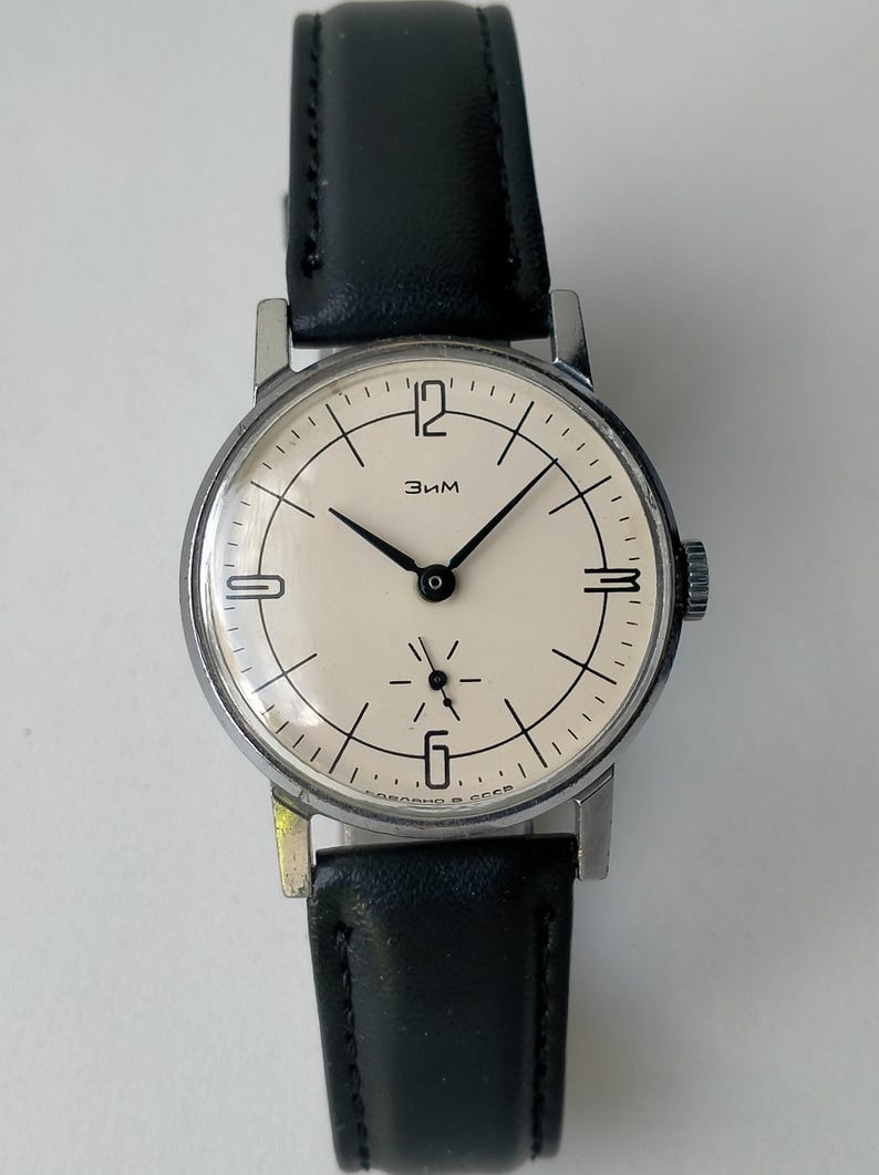 May include: A vintage wristwatch with a cream-colored face, black hands, and a black leather band. The watch has a silver-colored case and the word "Зим" is printed on the face. The watch band is approximately 0.75 inches wide.