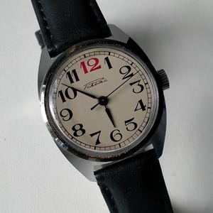 Peut inclure: Une montre-bracelet vintage avec un bracelet en cuir noir. Le cadran de la montre est de couleur crème avec des chiffres et des aiguilles noirs, et le chiffre 12 est rouge. La montre porte le mot "Takema" imprimé sur le cadran.