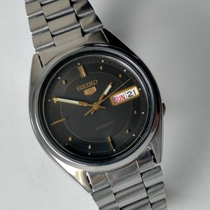 Op de afbeelding: Een zilverkleurig roestvrijstalen Seiko 5 automatisch polshorloge met een donkergrijze wijzerplaat, goudkleurige uurmarkeringen en goudkleurige wijzers. Het horloge heeft een dag-datumvenster op de drie-uurpositie met SUN 21 en een metalen schakelarmband.