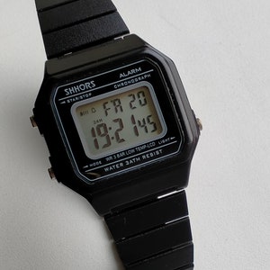 May include: Black digital wristwatch with a black resin band. The watch face displays the time, date, and day of the week. The watch is water resistant to 3 ATM.