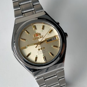 Può includere: Un orologio analogico in acciaio inossidabile color argento con quadrante color champagne, indici e lancette color oro. Il quadrante presenta il logo Orient, tre stelle, la scritta Crystal 21 JEWELS e una finestrella giorno-data che mostra SAB 11.