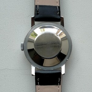 May include: A silver-toned wristwatch with a black leather band. The watch case back is visible, showing a circular design. The watch band is attached to the watch case with silver-toned metal bars. The watch band is black on the outside and light beige on the inside.