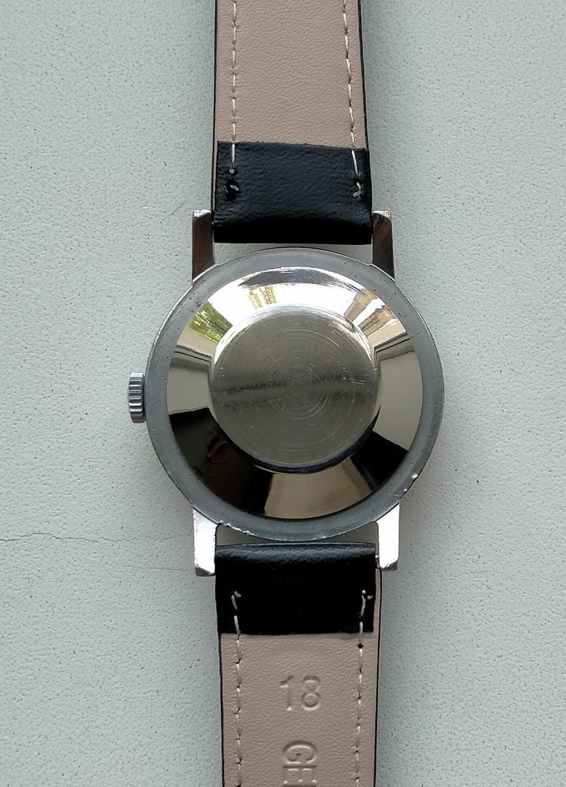 May include: A silver-toned wristwatch with a black leather band. The watch case back is visible, showing a circular design. The watch band is attached to the watch case with silver-toned metal bars. The watch band is black on the outside and light beige on the inside.