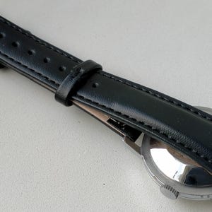 May include: A black leather watch strap with a silver buckle and a stainless steel watch case. The strap is stitched along the edges and has several holes for adjustment. The watch face is visible.