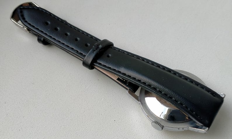 May include: A black leather watch strap with a silver buckle and a stainless steel watch case. The strap is stitched along the edges and has several holes for adjustment. The watch face is visible.
