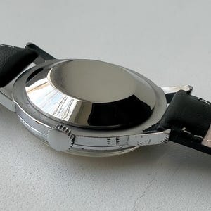 May include: A silver-toned wristwatch with a black leather band. The watch case is round and polished, reflecting light. The band is black on the outside and light beige on the inside, with "GENUINE LEATHER" stamped on it.