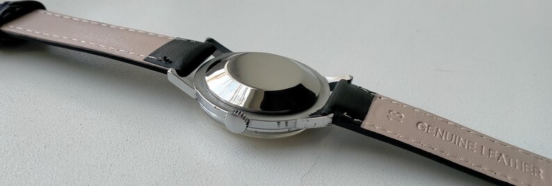 May include: A silver-toned wristwatch with a black leather band. The watch case is round and polished, reflecting light. The band is black on the outside and light beige on the inside, with "GENUINE LEATHER" stamped on it.