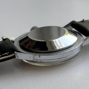 May include: A silver-toned wristwatch with a black leather strap. The watch case is circular and reflective, with a light-colored underside of the strap visible. The words "GENUINE LEATHER" are printed on the strap.