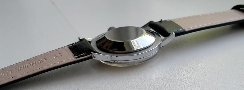 May include: A silver-toned wristwatch with a black leather strap. The watch case is circular and reflective, with a light-colored underside of the strap visible. The words "GENUINE LEATHER" are printed on the strap.