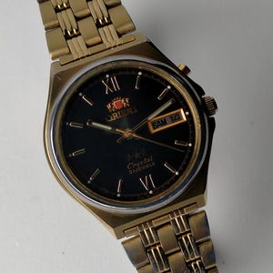 Könnte beinhalten: Goldfarbene Orient-Uhr mit schwarzem Zifferblatt. Die Uhr verfügt über römische Ziffern, eine Tagesdatumsanzeige und das Orient-Logo. Das Armband der Uhr ist in passendem Gold gehalten. Die Uhr ist ein klassischer Stil.