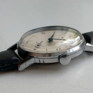 May include: A vintage wristwatch with a silver-colored case and a light-colored face. The watch has black hands and a black leather band. The watch is displayed on a white surface.