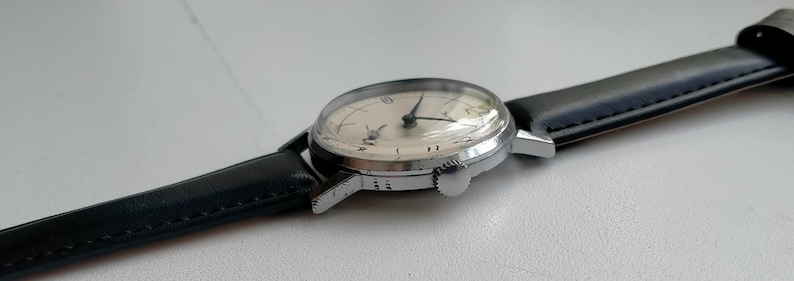 May include: A vintage wristwatch with a silver-colored case and a light-colored face. The watch has black hands and a black leather band. The watch is displayed on a white surface.